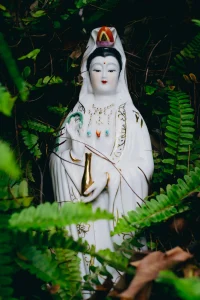 The Legend of Miao Shan: The Princess Who Became a Goddess A white porcelain statue of Guanyin holding a golden vase, nestled among lush green ferns.