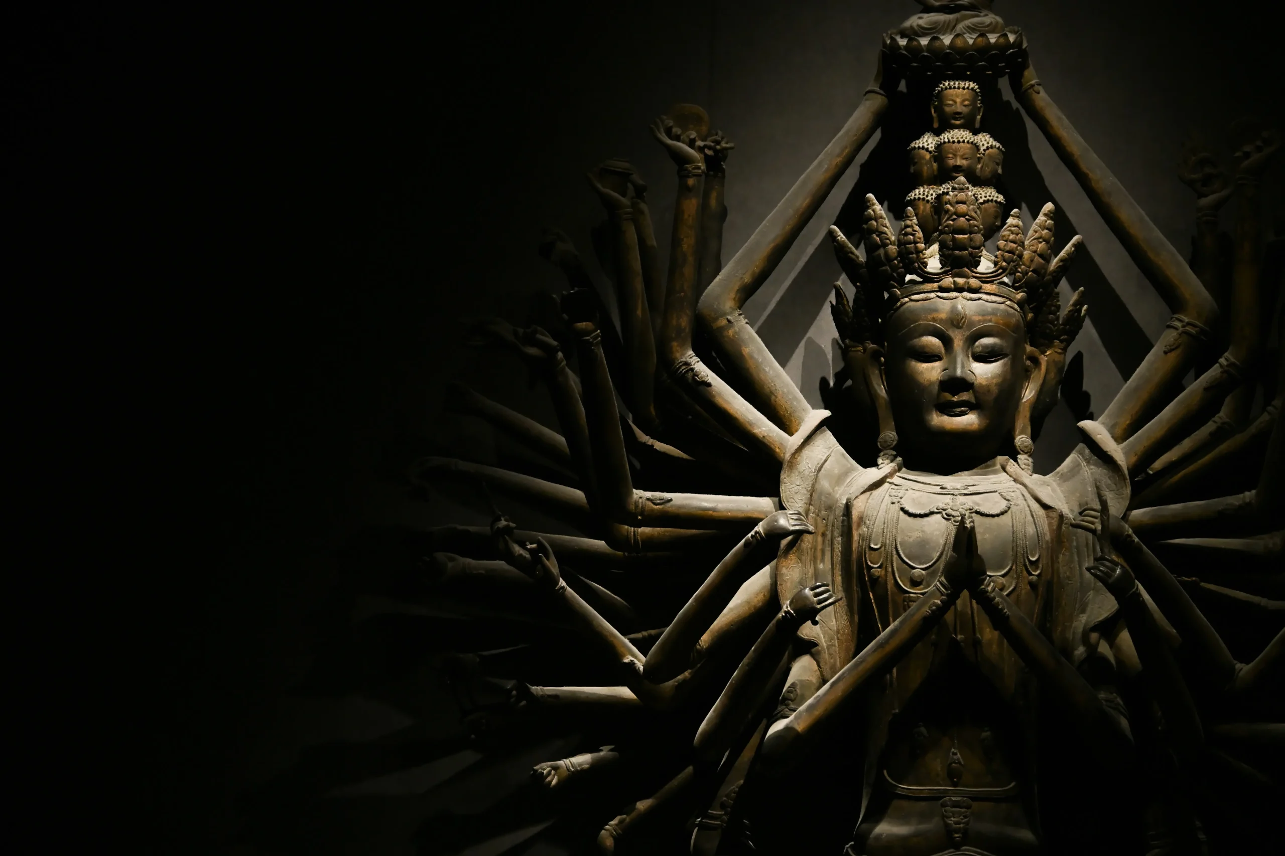 The Legend of Miao Shan: The Princess Who Became a Goddess A dramatic photograph of a multi-armed Buddhist statue, likely the Thousand-Armed Guanyin, illuminated against a pitch-black background. The figure features a serene expression, a tiered headdress, and numerous arms fanning out behind the body, with the central hands clasped in prayer.